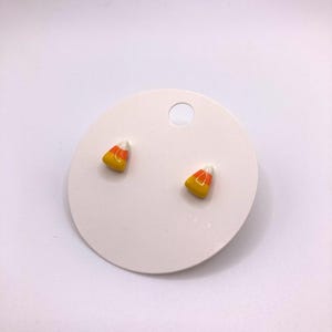 Könnte beinhalten: Ein Paar kleine Ohrringe in Form von Maisbonbons. Die Ohrringe sind aus weißem, gelbem und orangefarbenem Emaille.