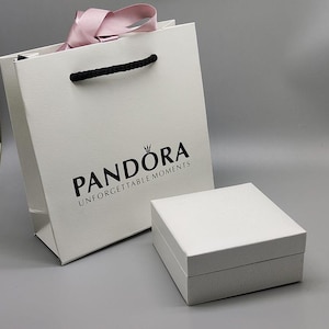 May include: A white gift bag and a square box. The bag features a black handle, a pink ribbon, and the brand name "PANDORA" printed in black. The box is white with a textured surface. The words "UNFORGETTABLE MOMENTS" are printed below the brand name.