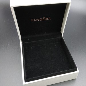 May include: White jewelry box with a black velvet interior. The box has the word "PANDORA" printed on the lid in gold lettering.