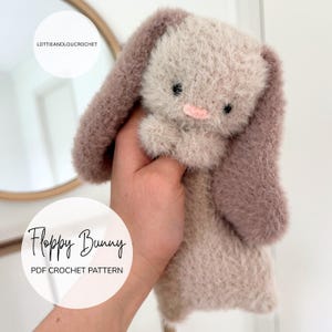 May include: A handmade, plush floppy bunny in soft, muted tones. The bunny features long, floppy ears in a dusty rose color, a light pink nose, and small black eyes. The text "Floppy Bunny PDF Crochet Pattern" is displayed on the image.