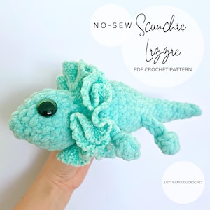 Peut inclure: Une peluche lézard turquoise au crochet, avec un grand œil vert et des volants. Le texte "NO-SEW Scunchie Lizzie PDF CROCHET PATTERN" est affiché dans l'image. La peluche est tenue dans une main.