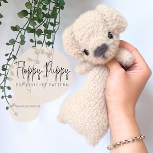 May include: A beige crochet puppy with floppy ears and a grey nose is held. The text "Floppy Puppy PDF Crochet Pattern" is visible. The puppy is a soft, plush toy. The item is handmade.