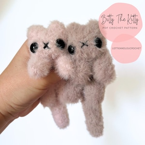 May include: Two small, fuzzy, pink cat-shaped crochet toys with black eyes and stitched details. The toys are held in a hand, with text that reads "Betty The Kitty PDF Crochet Pattern" and "lottieandloucrochet" in a pink circle.