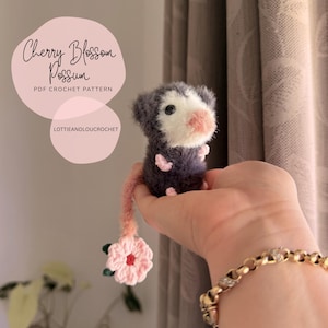 Könnte beinhalten: Ein handgehäkeltes Opossum mit einem rosa Kirschblütenschwanz. Das Opossum ist grau und weiß mit rosa Akzenten. Der Text "Cherry Blossom Possum PDF Crochet Pattern" befindet sich im Hintergrund.