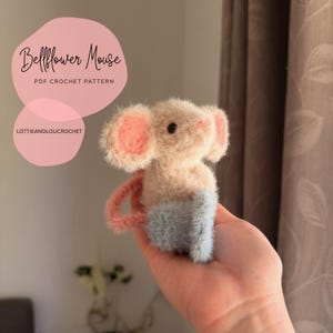 Peut inclure: Une souris au crochet faite à la main avec des oreilles roses, un corps rose clair et un bas bleu. La souris a un petit œil noir et une queue rose. Le texte "Bellflower Mouse PDF Crochet Pattern" est en arrière-plan.