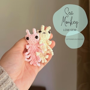 Könnte beinhalten: Zwei handgefertigte Sea Monkey-Figuren aus Häkelarbeit in Rosa und Creme. Die Figuren haben schwarze Augen und detaillierte Gliedmaßen. Das Bild enthält den Text "Sea Monkey" und "LOW-SEW" in einer hellblauen Blase, mit dem Shop-Namen "LOTTIEANDLOUCROCHET".