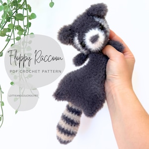 May include: A plush, gray and white raccoon toy with a striped tail. The text "Floppy Raccoon PDF Crochet Pattern" is displayed on the left side of the image. The raccoon is held in a hand against a white background.