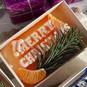 May include: A rectangular white gift box containing an orange-colored wax bar with the embossed white text MERRY CHRISTMAS. The bar is decorated with a realistic orange segment and a green pine needle sprig placed on top of the wax surface.