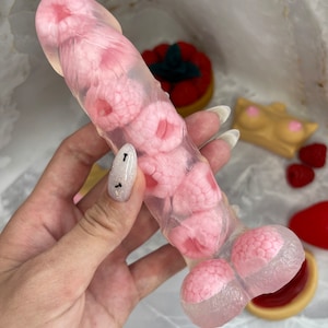 Puede incluir: Un objeto de forma fálica hecho de material transparente y translúcido con varios insertos en forma de frambuesa rosa incrustados en la estructura. El artículo tiene un cuerpo cilíndrico liso con una punta redondeada y una base más ancha.