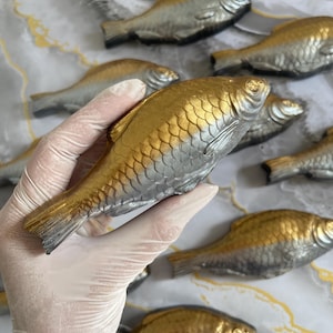 Könnte beinhalten: Dekorative Fischskulpturen mit metallischer Gold- und Silberoberfläche. Die Fische sind auf einer marmorierten Oberfläche angeordnet, einer wird in einer behandschuhten Hand gehalten. Die Fische haben detaillierte Schuppenmuster und Flossen.