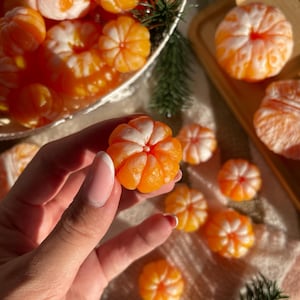 May include: Close-up of several peeled mandarin orange segments, with a hand holding one. The segments are bright orange with white sections, arranged in a decorative basket and on a wooden tray. A sprig of evergreen adds a festive touch.