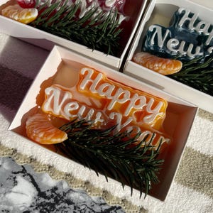 May include: A white rectangular gift box containing an orange-colored soap bar with the embossed white text Happy New Year, accompanied by a realistic orange segment and a small green pine branch, presented as a festive holiday gift set.