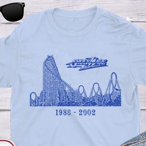 May include: Light blue t-shirt featuring a blue roller coaster graphic with the text "Shockwave" and the years "1988-2002". The shirt is displayed with sunglasses, jeans, and white sneakers.