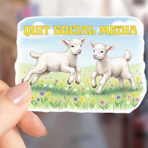 May include: A sticker featuring two white lambs running through a field of wildflowers under a blue sky. The text "QUIT SOCIAL MEDIA" is written across the top in yellow letters. The sticker is held by a person.