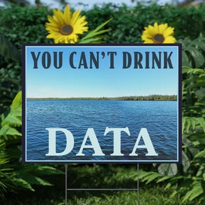 May include: A rectangular yard sign displaying the text "YOU CAN'T DRINK DATA" above a blue lake scene. The sign is placed in a green lawn, surrounded by sunflowers and verdant foliage.
