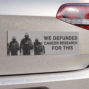 Può includere: Un adesivo rettangolare con la scritta "WE DEFUNDED CANCER RESEARCH FOR THIS" e un'immagine in scala di grigi di tre figure in tenuta tattica. L'adesivo è attaccato alla parte posteriore di un'auto argentata.