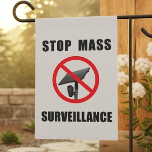May include: A white rectangular garden flag hanging on a black metal stand. The flag features the black text STOP MASS SURVEILLANCE and a red circle with a diagonal line through a black graphic of a solar-powered security camera.