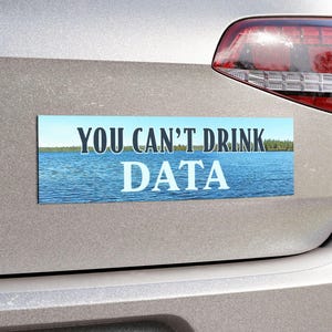 May include: A rectangular bumper sticker with the text "YOU CAN'T DRINK DATA" in white letters. The background of the sticker features a blue lake with a forest on the horizon. The sticker is affixed to the back of a silver car.