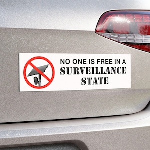 May include: A rectangular bumper sticker with a white background featuring a red circle with a diagonal line through a black security camera icon. The text reads NO ONE IS FREE IN A SURVEILLANCE STATE in bold black capital letters on the right side.