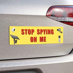 May include: A rectangular yellow bumper sticker with the red text STOP SPYING ON ME. The sticker features illustrations of a white security camera, a black solar panel on a stand, a grey security camera, and a vertical grey doorbell camera.