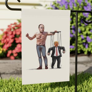 May include: A garden flag featuring an illustration of a shirtless man holding strings attached to a puppet in a suit. The puppet is a caricature of a man with orange hair. The background is a blurred outdoor scene with flowers and grass.