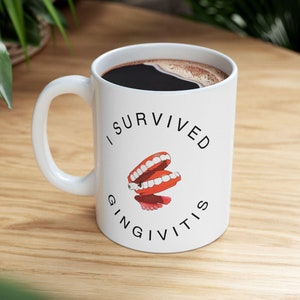 May include: White ceramic mug with a black coffee fill. The mug features the text "I SURVIVED GINGIVITIS" in black, with a graphic of cartoon teeth. The mug is on a wooden surface.