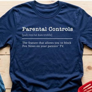 May include: A navy blue t-shirt with the text "Parental Controls" and a definition. The shirt is next to a pair of blue jeans, keys, a plant, and a red spatula on a wooden surface.