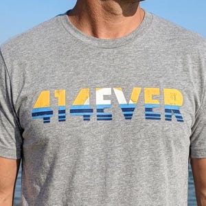 May include: A gray crew neck t-shirt with the word "414EVER" printed across the chest in yellow, white, and blue. The shirt is short-sleeved and made of a soft material. The background shows a beach and the ocean.