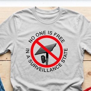 May include: A light gray short-sleeved t-shirt featuring a graphic of a security camera inside a red prohibition circle. The text "NO ONE IS FREE IN A SURVEILLANCE STATE" is printed in black capital letters around the circular red border.