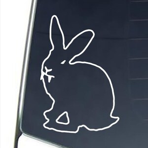 May include: A white vinyl decal of a rabbit silhouette with prominent fangs, applied to a dark glass surface. The design features the outline of a seated bunny with long ears and sharp teeth visible in the mouth area, creating a stark contrast.