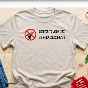 May include: A beige short-sleeved t-shirt featuring a graphic of a black satellite dish inside a red prohibition circle, with the black text "DEFLOCK AMERICA" printed to the right of the icon. The shirt is displayed alongside blue jeans.