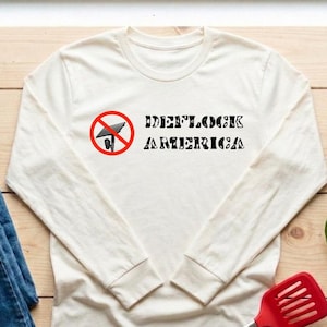 May include: A long-sleeve cream-colored shirt featuring a graphic of a red circle with a diagonal line through a black security camera icon, followed by the black text "DEFLOCK AMERICA" in a stylized, distressed font across the chest.