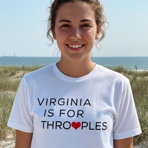 May include: White t-shirt with the text "VIRGINIA IS FOR THRO PLES" in black, with a red heart replacing the "O". The shirt is worn by a person on a beach with sunglasses on their head.