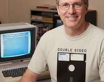 Puede incluir: Un hombre con gafas y una camiseta beige con un gráfico de disquete negro y las palabras "DOUBLE SIDED DOUBLE DENSITY". Está sentado frente a una computadora vintage con un monitor que muestra texto.