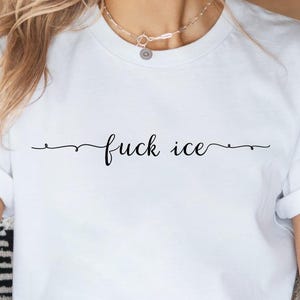 May include: White t-shirt with the phrase "fuck ice" in a cursive font. The text is centered and underlined with a thin, wavy line. The shirt has short sleeves and a crew neck.