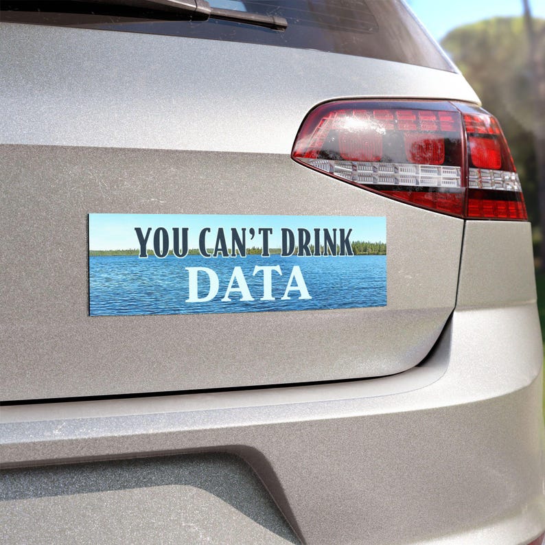 May include: A rectangular bumper sticker with the text "YOU CAN'T DRINK DATA" in white letters. The background of the sticker features a blue lake with a forest on the horizon. The sticker is affixed to the back of a silver car.