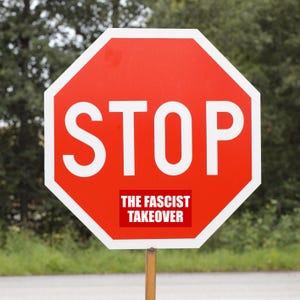 May include: A red octagonal stop sign with the word "STOP" in large white capital letters. A smaller rectangular sign below reads "THE FASCIST TAKEOVER". The sign is mounted on a metal pole against a blurred green background.
