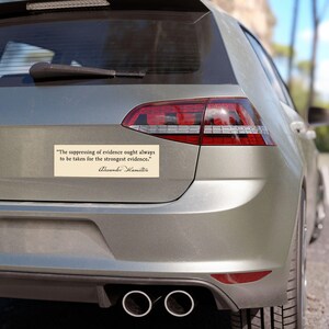 May include: A rectangular bumper sticker with a quote by Alexander Hamilton, in black text on a cream background, is affixed to the rear of a silver car. The quote reads, "The suppressing of evidence ought always to be taken for the strongest evidence."