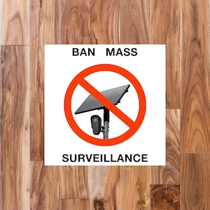 May include: A square white sign featuring the black text BAN MASS at the top and SURVEILLANCE at the bottom. In the center, a large red circle with a diagonal line overlays a graphic of a black solar-powered security camera on a metal pole.