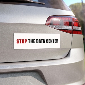 May include: A white rectangular bumper sticker with the text "STOP THE DATA CENTER" in red and black lettering is affixed to the rear of a silver car. The car's taillight is visible.