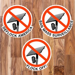 May include: Three circular stickers featuring a red prohibition symbol over a black security camera with a solar panel. The stickers display the text DEFLOCK AMERICA, BAN MASS SURVEILLANCE, and FLOCK OFF! in bold black capital letters along the bottom.