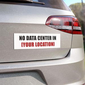 May include: A white rectangular bumper sticker with black text that reads "NO DATA CENTER IN [YOUR LOCATION]" is affixed to the back of a silver car. The text is in a bold, sans-serif font. The words "YOUR LOCATION" are in red.