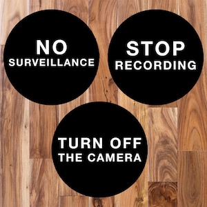 May include: Three circular black signs with white text. The top left sign reads NO SURVEILLANCE. The top right sign reads STOP RECORDING. The bottom center sign reads TURN OFF THE CAMERA. These signs feature bold, sans-serif lettering on a solid background.