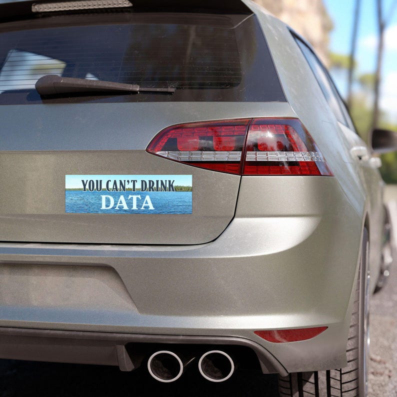 May include: A rectangular bumper sticker on the back of a silver car. The sticker has a blue and white background with the text "YOU CAN'T DRINK DATA" in white capital letters. The car's taillight is red.