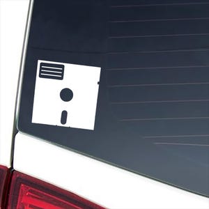 May include: A white vinyl decal in the shape of a classic 3.5-inch floppy disk. The design features the iconic square silhouette with a rectangular label area at the top, a circular center hub, and a vertical slot at the bottom edge of the disk.