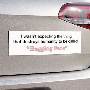 May include: A rectangular white bumper sticker with black text that reads: I wasn't expecting the thing that destroys humanity to be called "Hugging Face". The words "Hugging Face" are written in a light pink font with a thin black outline.
