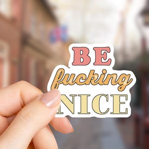 May include: A die-cut sticker featuring the text BE in pink, fucking in a textured orange, and NICE in pale yellow. The letters have a thin black outline and a white border, creating a bold, layered appearance with distinct typography styles.