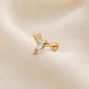 May include: A gold-colored hummingbird stud earring with a clear, teardrop-shaped gemstone in its body. The earring has a flat back and a post for piercing. The hummingbird is depicted in flight, with its wings spread.
