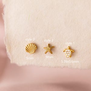 Puede incluir: Tres pendientes dorados con temática de playa. Los estilos incluyen una concha (9 mm), una estrella de mar (9 mm) y una tortuga (5,76x5,6 mm). Los pendientes se muestran sobre una superficie clara.