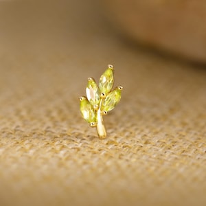 Op de afbeelding: Een goudkleurige oorbel in de vorm van een klein blad. Het blad heeft vijf lichtgroene, marquise-geslepen edelstenen. De oorbel is geplaatst tegen een neutrale, getextureerde achtergrond.